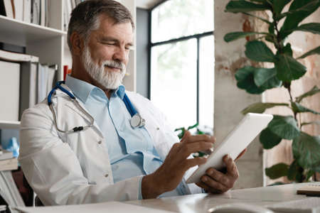 Cheerful mature doctor using digital tablet in clinic.