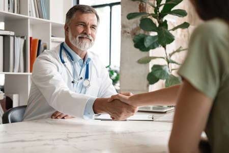Cheerful mature doctor shaking hands to patient in the office at deskの写真素材