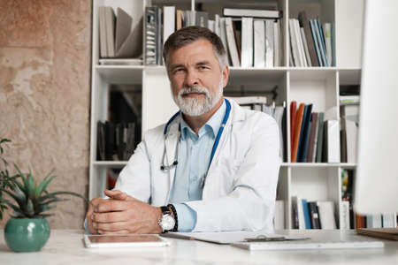 Portrait of handsome mature doctor sitting at desk in modern office.