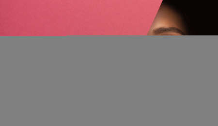 Face of a young beautiful girl with a bright make-up and with plump red lips peeks into a hole in pink paper.の写真素材