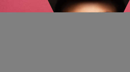 Face of a young beautiful girl with a bright make-up and with plump red lips peeks into a hole in pink paper.の写真素材