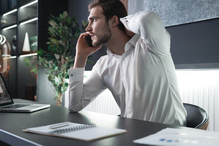 35s businessman in suit sit at workplace in modern office talk on smartphone, having business phonecall, provide help to client. Lifestyle, workflow conceptの写真素材