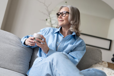 Happy beautiful relaxed mature gray-haired woman drinking coffee relaxing on sofa at home. Smiling stylish middle aged 50s lady enjoying resting sitting on couch in modern living room.の写真素材