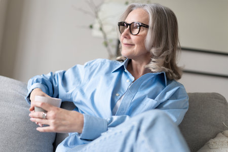 Happy beautiful relaxed mature gray-haired woman drinking coffee relaxing on sofa at home. Smiling...