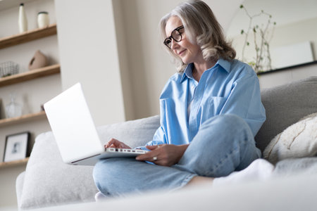 Smiling 50s mature woman sitting at home on sofa, holding laptop. Senior happy business woman using...
