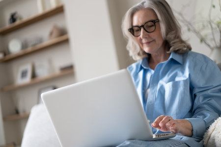 Smiling 50s mature woman sitting at home on sofa, holding laptop. Senior happy business woman using pc technologies, working, chatting, spending time in social media internet, ecommerce.の写真素材