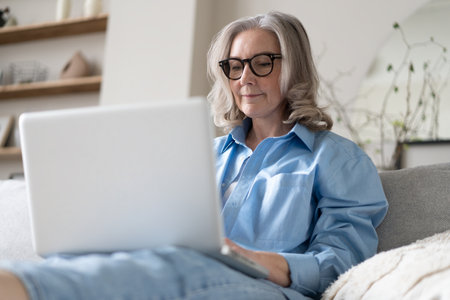 Smiling 50s mature woman sitting at home on sofa, holding laptop. Senior happy business woman using pc technologies, working, chatting, spending time in social media internet, ecommerce.の写真素材