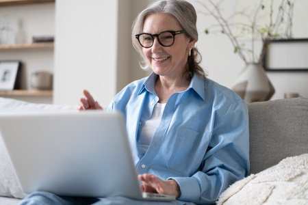 Smiling 50s mature woman sitting at home on sofa, holding laptop. Senior happy business woman using pc technologies, working, chatting, spending time in social media internet, ecommerce.の写真素材