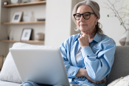 Smiling 50s mature woman sitting at home on sofa, holding laptop. Senior happy business woman using pc technologies, working, chatting, spending time in social media internet, ecommerce.の写真素材