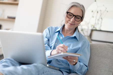 Smiling 50s mature woman sitting at home on sofa, holding laptop. Senior happy business woman using pc technologies, working, chatting, spending time in social media internet, ecommerce.の写真素材