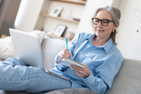 Smiling 50s mature woman sitting at home on sofa, holding laptop. Senior happy business woman using pc technologies, working, chatting, spending time in social media internet, ecommerce.の写真素材
