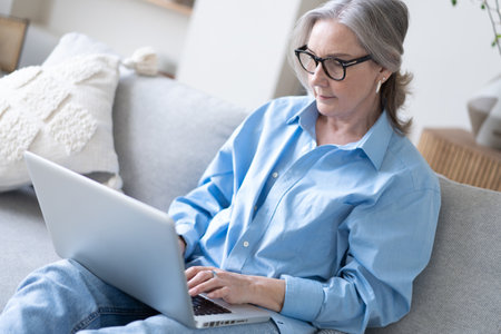 Smiling 50s mature woman sitting at home on sofa, holding laptop. Senior happy business woman using pc technologies, working, chatting, spending time in social media internet, ecommerce.の写真素材