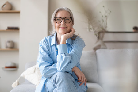 Happy mature woman relaxing on her couch at home in the living room. senior woman looking at camera. Portrait of happy woman in blue shirt smiling.の写真素材