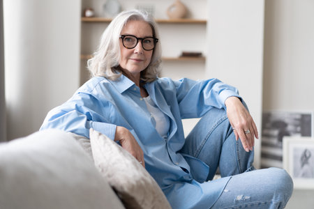 Happy mature woman relaxing on her couch at home in the living room. senior woman looking at camera. Portrait of happy woman in blue shirt smiling.の写真素材