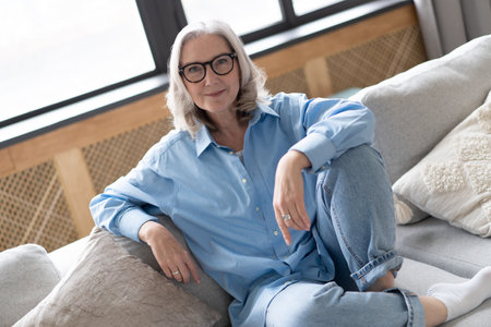 Happy mature woman relaxing on her couch at home in the living room. senior woman looking at camera. Portrait of happy woman in blue shirt smiling.の写真素材