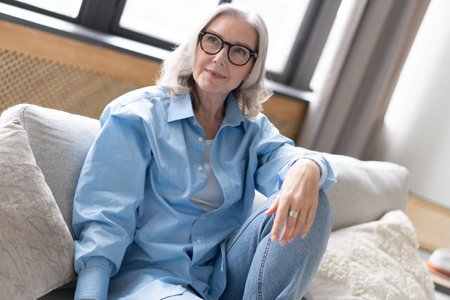 Happy mature woman relaxing on her couch at home in the living room. Portrait of happy woman in blue shirt smiling.の写真素材