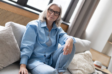 Happy mature woman relaxing on her couch at home in the living room. Portrait of happy woman in blue shirt smiling.の写真素材