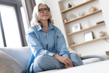 Happy mature woman relaxing on her couch at home in the living room. Portrait of happy woman in blue shirt smiling.の写真素材