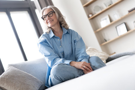 Happy mature woman relaxing on her couch at home in the living room. Portrait of happy woman in blue shirt smiling.の写真素材