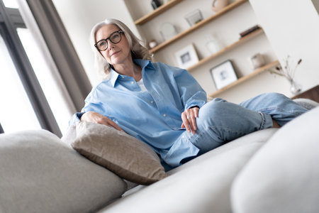 Happy mature woman relaxing on her couch at home in the living room. Portrait of happy woman in blue shirt smiling.の写真素材