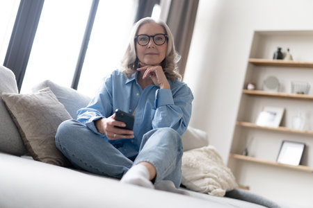 Pleasant mature lady resting on couch, using applications on smartphone. Happy old woman chatting in messenger or social network with friends or children, shopping in internet store.の写真素材