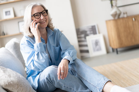 Happy pretty mature woman speaking on mobile phone portrait. Senior gray haired lady making telephone call, sitting on couch at home, enjoying talk, communication, smilingの写真素材