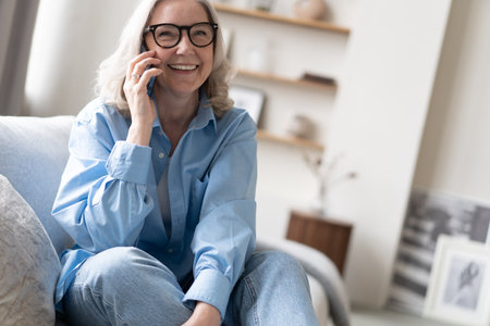Happy pretty mature woman speaking on mobile phone portrait. Senior gray haired lady making telephone call, sitting on couch at home, enjoying talk, communication, smilingの写真素材