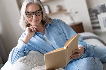 Happy older gray haired woman sitting on comfortable sofa in living room, reading interesting book, enjoying leisure weekend time with favorite novel alone at home.の写真素材