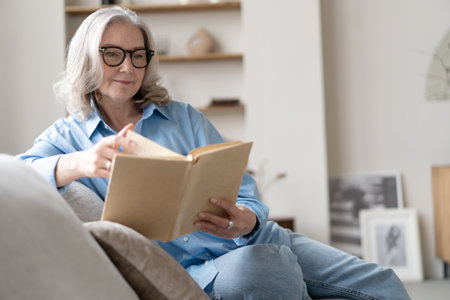 Happy older gray haired woman sitting on comfortable sofa in living room, reading interesting book, enjoying leisure weekend time with favorite novel alone at home.の写真素材