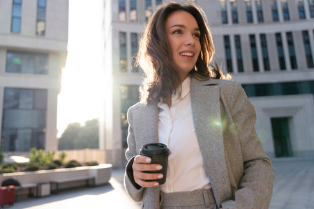 Beautiful Woman Going To Work With Coffee Standing Near Office Building. Portrait Of Successful Business Woman Holding Cup Of Hot Drinkの写真素材