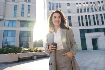 Beautiful Woman Going To Work With Coffee Standing Near Office Building. Portrait Of Successful Business Woman Holding Cup Of Hot Drinkの写真素材