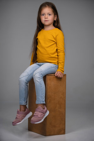 Full body of smiling little girl in sweatshirt and jeans sitting on box in studio against white backgroundの写真素材