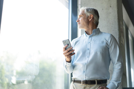 Smiling happy confident mid aged male company ceo executive holding cellphone standing in office...
