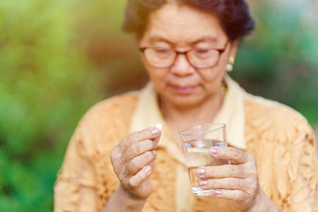 An old Thai woman sits and looks at the pill in her hand with a glass of water to eat and relieve sickness. Health care concept.の写真素材