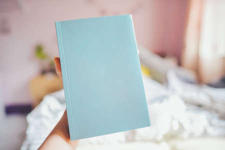 Hand holding a blue book with a blank cover, there is room for text in the bedroom.の写真素材