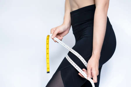 A skinny woman wearing black pants uses a tape measure around her thigh against with gray...