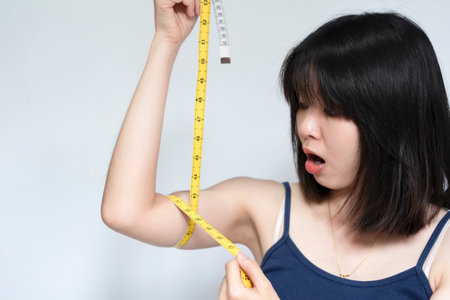 An Asian woman measures her arm with a tape measure and poses in shock at the result. Beauty or health concept.の写真素材