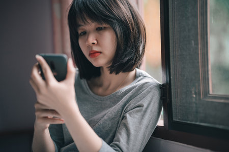 A woman sitting by the bedroom window using her phone with a indifferent expression on her face.