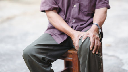 elderly man sits on wooden stool, resting his hand on his knee, conveying sense of contemplation and calmness. His attire is casual, featuring short sleeved shirt and loose fitting pants, reflecting.の写真素材