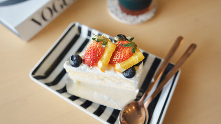 A delicious slice of cake topped with fresh strawberries, blueberries, and citrus, served on a striped plate with spoons beside it.の写真素材