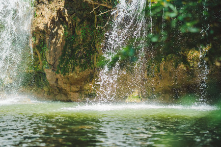 A serene waterfall cascades over a rocky surface, creating a misty spray above a calm green pool, surrounded by lush greenery, in Tak Thailand.の写真素材
