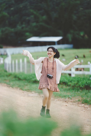 A joyful girl in a checked dress walks along a country road, wearing a camera, surrounded by lush greenery and a farm setting.の写真素材
