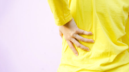 A person in a yellow shirt is holding their lower back, suggesting discomfort or pain, against a soft pink background.の写真素材