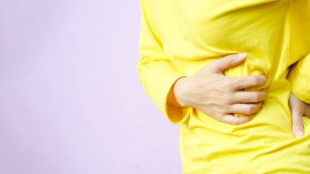 A person in a yellow shirt holds their stomach, suggesting discomfort or pain, set against a soft pink background.の写真素材