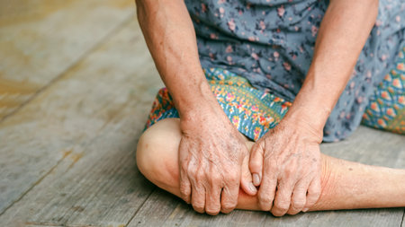 Elderly person sitting on wooden floor holding leg, showing pain or discomfort, wearing patterned clothing, concept of joint pain, aging, health issues, and senior care.の写真素材