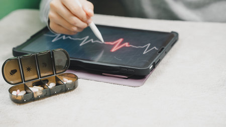 Close up of a digital tablet displaying an ECG waveform beside a weekly pill organizer, illustrating remote health monitoring, telemedicine, medication management, cardiac care, and digital healthcare.