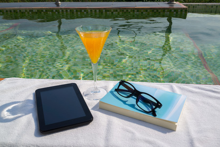 Tablet with empty screen, a glass of orange juice and blue book with glasses on the white towel next to the swimming pool with ripples and green mosaic tile bottom.の写真素材