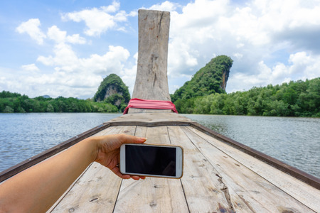 Female hand holding phone in boat with mountain, river and sky background.の写真素材