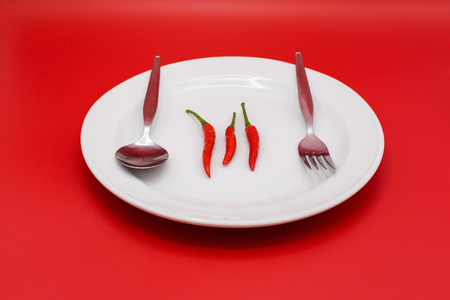 Three red chilis on white plate with cutlery and red background.の写真素材