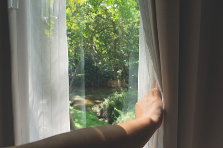 Asian female hand opening white curtains with green forest and river view background.